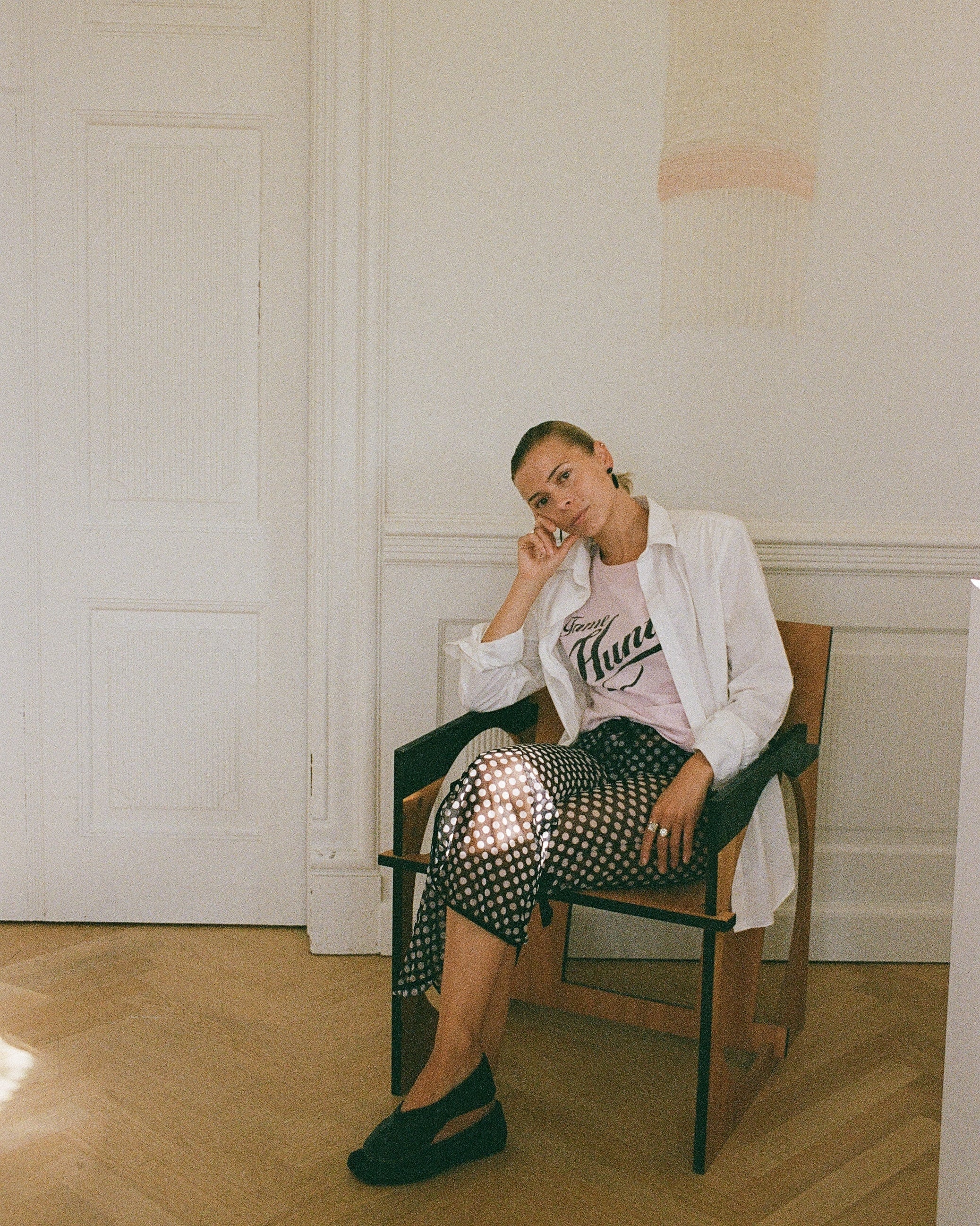 A woman sits in a wooden chair in a bright, minimalist room with white walls and light wood floors. She is wearing a white button-up shirt over a pink graphic t-shirt, a black-and-white patterned skirt, and black wedge shoes.